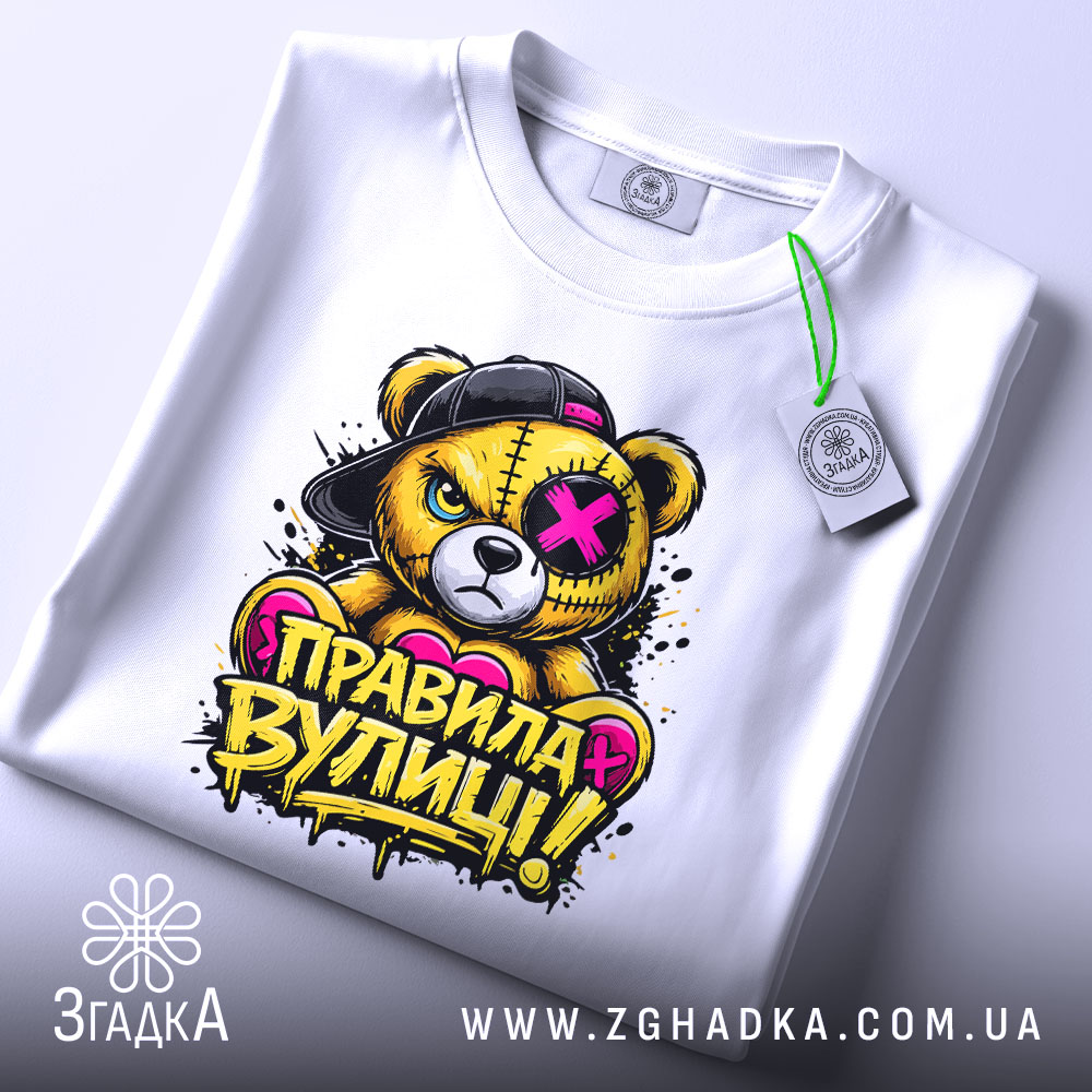 Футболка правила вулиці, біла, зображена зверху на плоскій поверхні, фон світло-сірий, принт з ведмедем у кепці, напис 'ПРАВИЛА ВУЛИЦІ!' яскравими кольорами, на футболці є етикетка з зеленим шнурком.