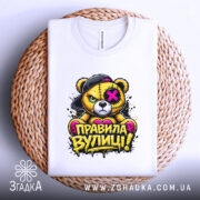 Футболка правила вулиці, біла, зображена зверху на плоскій поверхні, фон світло-сірий, принт з ведмедем у кепці, напис 'ПРАВИЛА ВУЛИЦІ!' яскравими кольорами, футболка лежить на плетеній основі.