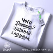 Футболка про обійми, білого кольору, складена на поверхні, фон світло-сірий, принт чорного кольору з текстом 'Чого Дивишся? Обіймай і наливай.', на комірці видно етикетку.