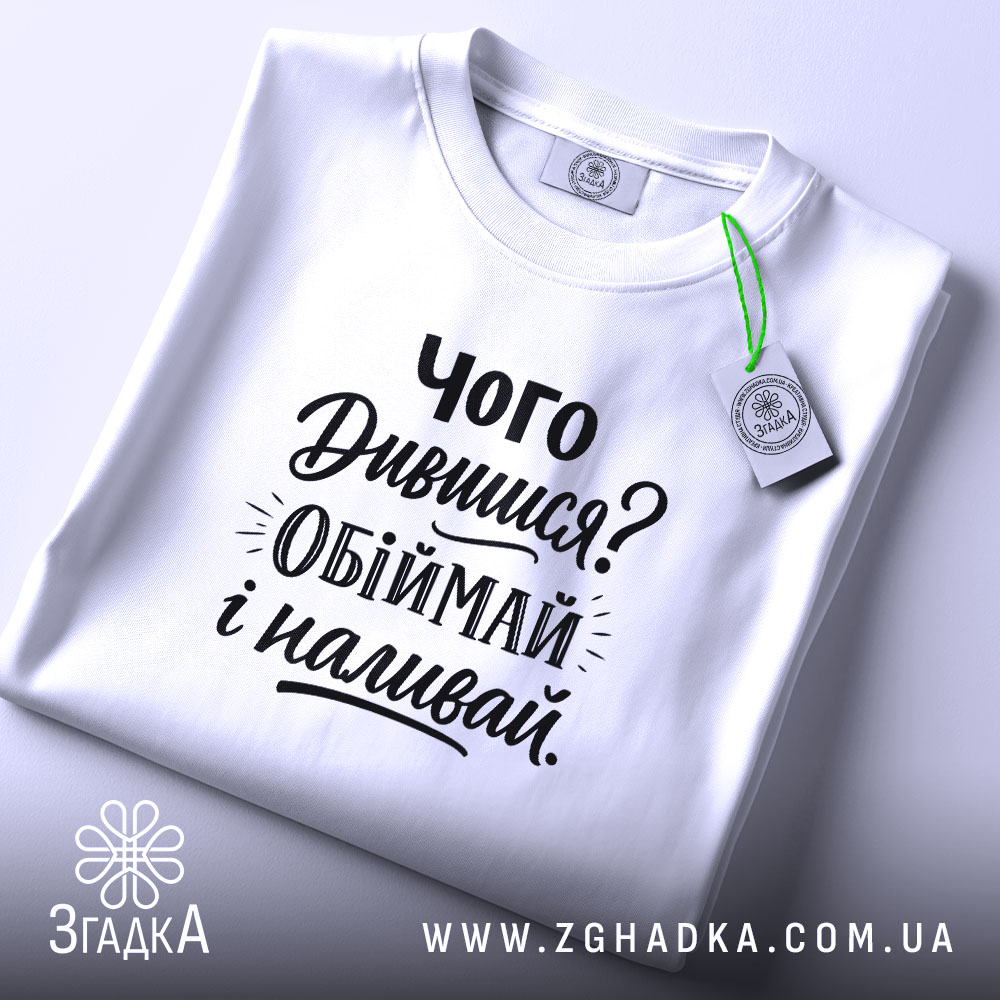 Футболка про обійми, білого кольору, складена на поверхні, фон світло-сірий, принт чорного кольору з текстом 'Чого Дивишся? Обіймай і наливай.', на комірці видно етикетку.