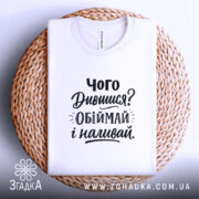 Футболка про обійми, білого кольору, складена на плетеній кошику, фон світло-сірий, принт чорного кольору з текстом 'Чого Дивишся? Обіймай і наливай.'