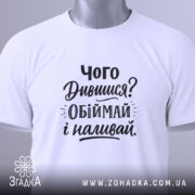 Футболка про обійми, білого кольору, складена на поверхні, фон світло-сірий, принт чорного кольору з текстом 'Чого Дивишся? Обіймай і наливай.', на комірці видно етикетку.