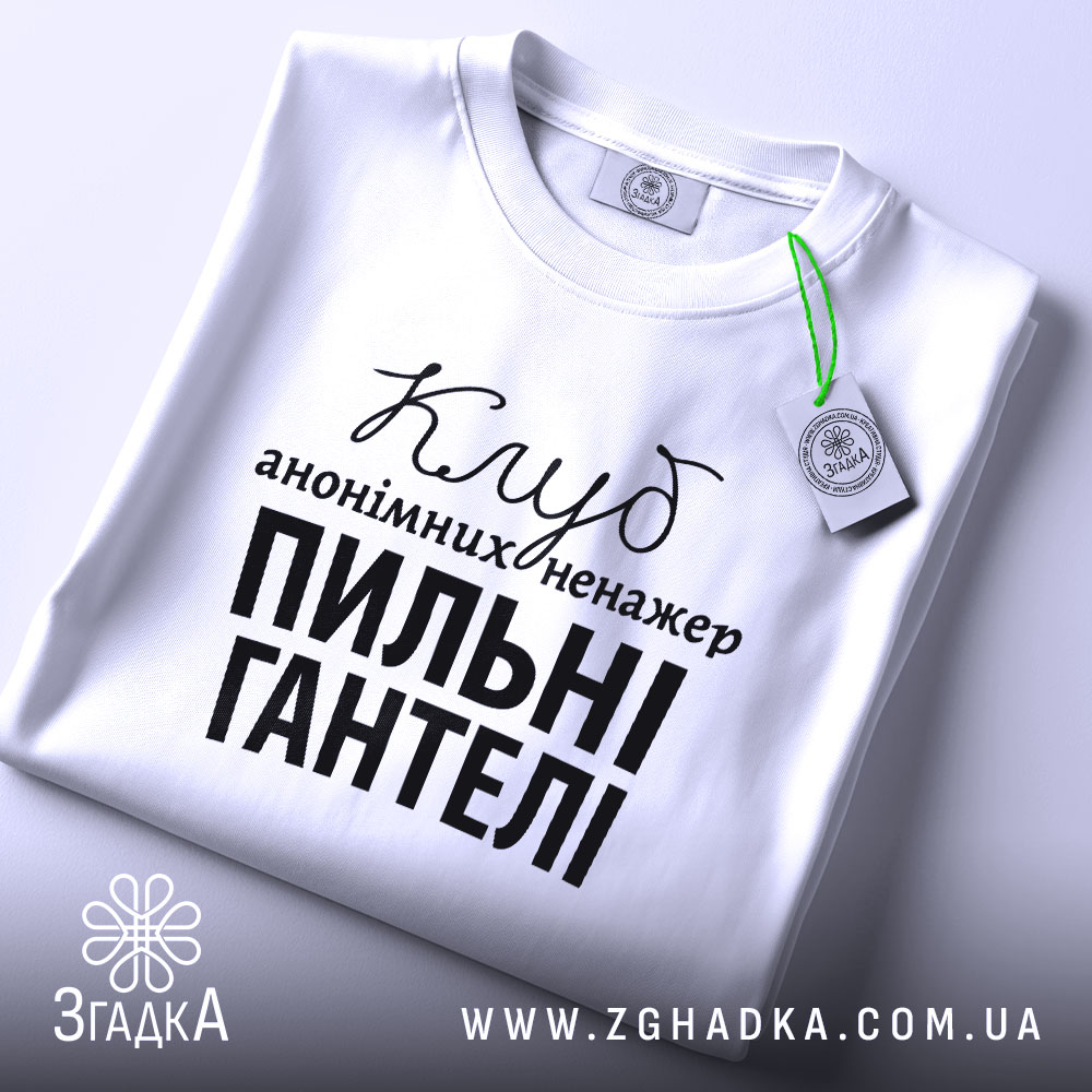 Футболка пильні гантелі, біла, зображена зверху, на столі, фон світло-сірий, принт чорного кольору з текстом 'Клуб анонімних менеджерів' та 'Пильні гантелі', з зеленим ярликом.