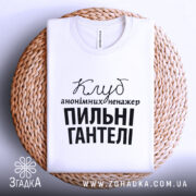 Футболка пильні гантелі, біла, зображена на плетеній тарілці, фон світло-сірий, принт чорного кольору з текстом 'Клуб анонімних менеджерів' та 'Пильні гантелі'.