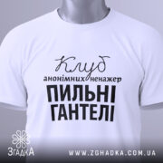 Футболка пильні гантелі, біла, зображена зверху, на столі, фон світло-сірий, принт чорного кольору з текстом 'Клуб анонімних менеджерів' та 'Пильні гантелі', з зеленим ярликом.