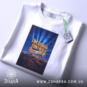 Футболка Пиздьож Пікчерз білого кольору, складена на столі, фон світло-сірий, принт з написом на синьому фоні з променями, зображення гір на нижній частині принта, на комірці прикріплена етикетка.