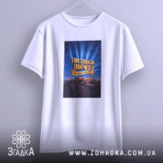 Футболка Пиздьож Пікчерз білого кольору, підвішена на вішалці, фон світло-сірий, принт з написом на синьому фоні з променями, зображення гір на нижній частині принта.