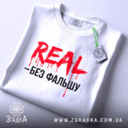 Футболка REAL без фальшу, білого кольору, складена на поверхні, фон світло-сірий, принт червоний з чорним текстом, на футболці є етикетка з зеленим шнурком, що додає акценту.
