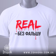 Футболка REAL без фальшу, білого кольору, складена на поверхні, фон світло-сірий, принт червоний з чорним текстом, на футболці є етикетка з зеленим шнурком, що додає акценту.