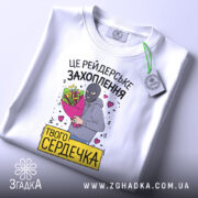 Футболка рейдерське захоплення, біла, складена на поверхні, фон світло-сірий, принт з малюнком людини в масці з букетом квітів та написом, з етикеткою.