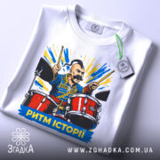 Футболка ритм історії, біла, складена на столі, фон світло-сірий, принт з чоловіком на барабанах, яскраві кольори, текст "РИТМ ІСТОРІЇ" внизу, на футболці є етикетка.
