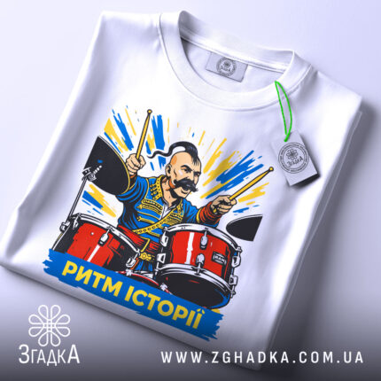 Футболка ритм історії, біла, складена на столі, фон світло-сірий, принт з чоловіком на барабанах, яскраві кольори, текст "РИТМ ІСТОРІЇ" внизу, на футболці є етикетка.