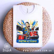 Футболка ритм історії, біла, складена на плетеній кошику, фон білий, принт з чоловіком на барабанах, яскраві кольори, текст "РИТМ ІСТОРІЇ" внизу.