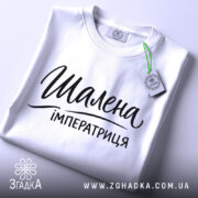 Футболка шалена імператриця, білого кольору, складена на поверхні з верхнього ракурсу, фон світлий, принт чорного кольору з написом "Шалена імператриця" в елегантному стилі, з зеленим ярликом.