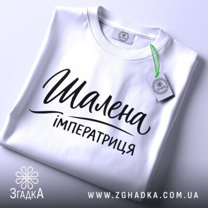 Футболка шалена імператриця, білого кольору, складена на поверхні з верхнього ракурсу, фон світлий, принт чорного кольору з написом "Шалена імператриця" в елегантному стилі, з зеленим ярликом.