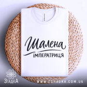 Футболка шалена імператриця, білого кольору, складена на плетеній основі з верхнього ракурсу, фон світлий, принт чорного кольору з написом "Шалена імператриця" в стильному шрифті.