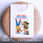 Футболка шалена подруга завжди білого кольору, зображена зверху на плетеному фоні. Принт містить двох дівчат, одна з яких стоїть на руках, а інша тримає келих, оточені кольоровими зірочками та пляшками.