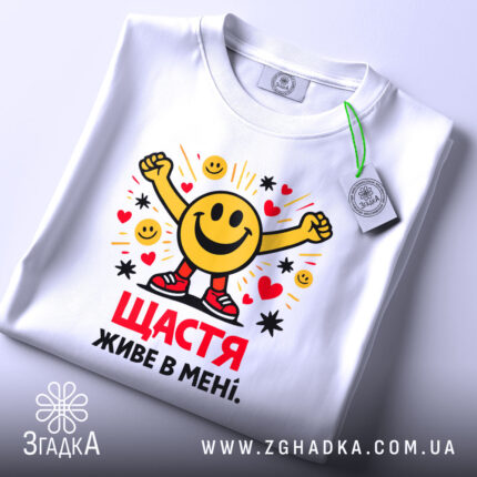Футболка щастя живе в мені. Біла футболка, складена на столі, з принтом усміхненого смайлика, що піднімає руки. На футболці є етикетка з зеленим шнурком. Принт яскравий, з сердечками та зірочками.