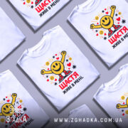 Футболка щастя живе в мені. Біла футболка на манекені, з яскравим принтом у вигляді усміхненого смайлика з піднятими руками, оточеного сердечками та зірочками. Текст на футболці червоний, розташований під принтом.