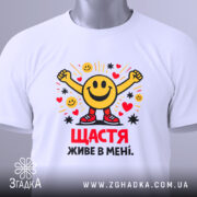 Футболка щастя живе в мені. Біла футболка, складена на столі, з принтом усміхненого смайлика, що піднімає руки. На футболці є етикетка з зеленим шнурком. Принт яскравий, з сердечками та зірочками.