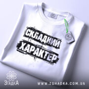 Футболка складний характер, біла, складена на поверхні, фон світло-сірий, принт чорний з текстом 'СКЛАДНИЙ ХАРАКТЕР', з етикеткою зеленого кольору.