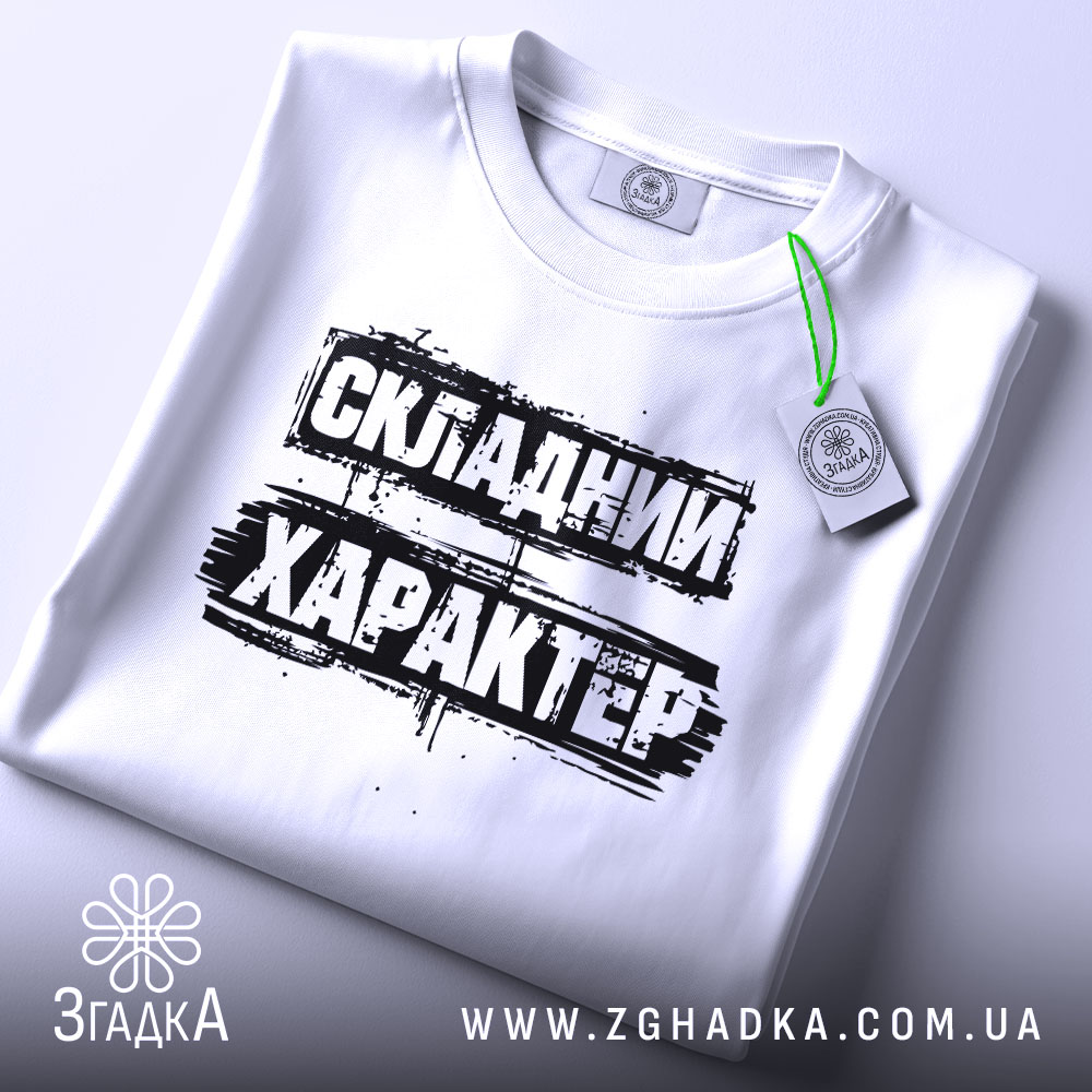 Футболка складний характер, біла, складена на поверхні, фон світло-сірий, принт чорний з текстом 'СКЛАДНИЙ ХАРАКТЕР', з етикеткою зеленого кольору.