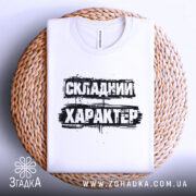 Футболка складний характер, біла, складена на плетеній кошику, фон світло-сірий, принт чорний з текстом 'СКЛАДНИЙ ХАРАКТЕР'.