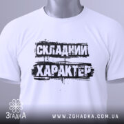 Футболка складний характер, біла, складена на поверхні, фон світло-сірий, принт чорний з текстом 'СКЛАДНИЙ ХАРАКТЕР', з етикеткою зеленого кольору.