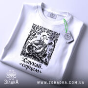 Футболка слухай серцем, біла, складена на поверхні, фон світло-сірий, принт чорного кольору з зображенням чоловіка, що грає на бандурі, з етикеткою зеленого кольору.