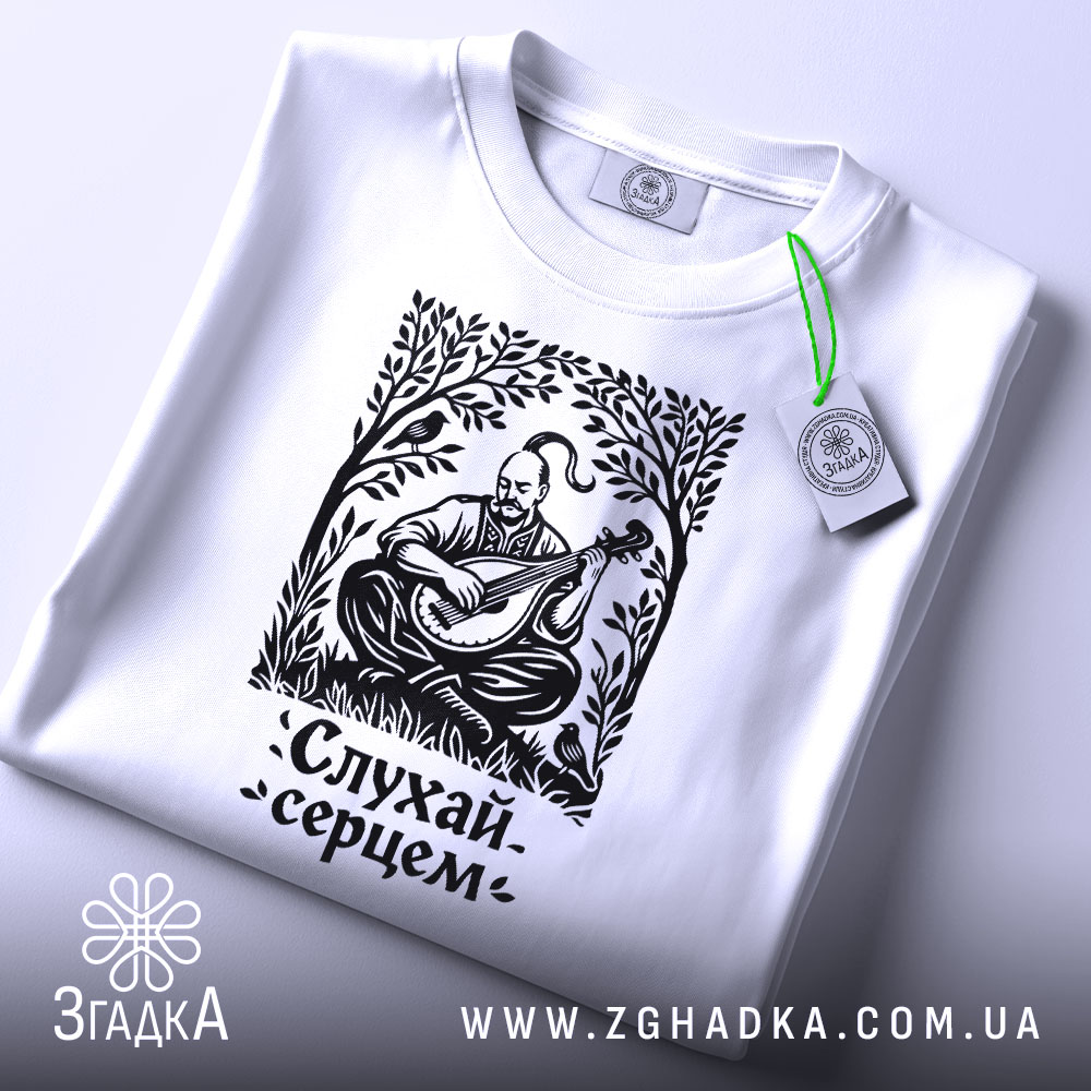 Футболка слухай серцем, біла, складена на поверхні, фон світло-сірий, принт чорного кольору з зображенням чоловіка, що грає на бандурі, з етикеткою зеленого кольору.