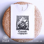 Футболка слухай серцем, біла, складена на плетеній кошику, фон світло-сірий, принт чорного кольору з зображенням чоловіка, що грає на бандурі, оточений рослинністю.