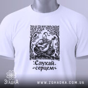 Футболка слухай серцем, біла, складена на поверхні, фон світло-сірий, принт чорного кольору з зображенням чоловіка, що грає на бандурі, з етикеткою зеленого кольору.