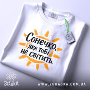 Футболка сонечко не світить. Біла футболка, складена на столі, з видимим етикеткою, фон світло-сірий, принт з написом "Сонечко, яке тобі не світить" оранжевого кольору з жовтими променями.