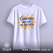 Футболка сонечко не світить. Біла футболка з коротким рукавом, підвішена на вішалці, фон світло-сірий, принт з написом "Сонечко, яке тобі не світить" оранжевого кольору з жовтими променями.