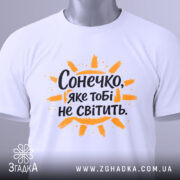 Футболка сонечко не світить. Біла футболка, складена на столі, з видимим етикеткою, фон світло-сірий, принт з написом "Сонечко, яке тобі не світить" оранжевого кольору з жовтими променями.