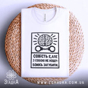 Футболка совість є білого кольору, складена на плетеній кошику, фон світло-сірий, принт чорного кольору з малюнком мозку та наручниками, текст українською мовою.
