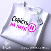 Футболка совість на паузі, білого кольору, складена на поверхні, фон світло-сірий, принт чорного та рожевого кольорів з написом 'Совість на паузі' і символом паузи, з етикеткою на рукаві.