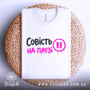 Футболка совість на паузі, білого кольору, складена на плетеній кошику, фон білий, принт чорного та рожевого кольорів з написом 'Совість на паузі' і символом паузи.