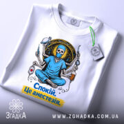 Футболка спокій це анестезія, біла, зображена з верхнього ракурсу, фон світлий, принт з черепом у медичному одязі, що сидить у позі лотоса, з медичними інструментами, текст на футболці жовтого кольору, на комірці є етикетка.