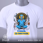 Футболка спокій це анестезія, біла, зображена з верхнього ракурсу, фон світлий, принт з черепом у медичному одязі, що сидить у позі лотоса, з медичними інструментами, текст на футболці жовтого кольору, на комірці є етикетка.