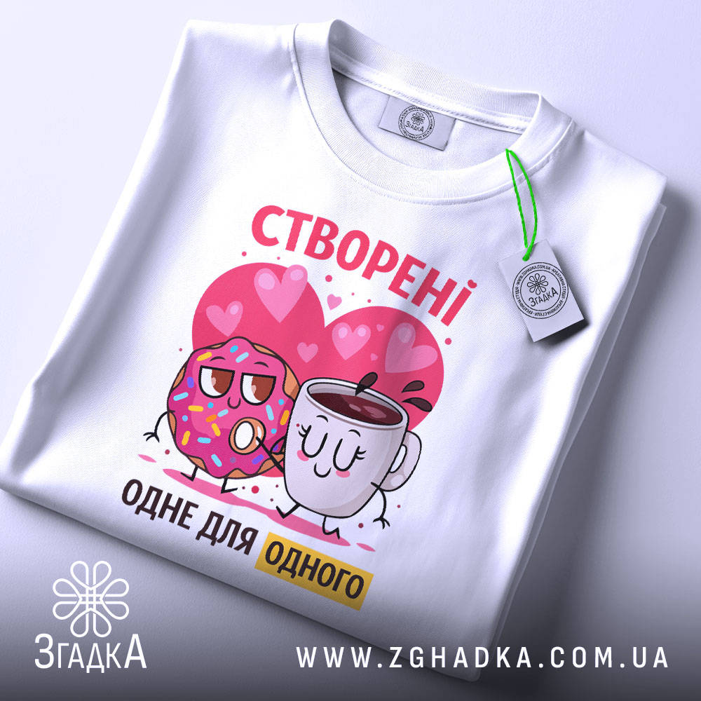 Футболка створені одне для одного. Біла футболка, складена на столі, з принтом, де пончик і чашка з кавою обіймаються на фоні сердечок. Яскраві кольори принта, видно етикетку з логотипом.