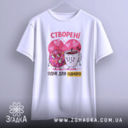 Футболка створені одне для одного. Біла футболка, підвішена на вішалці, з принтом, що зображує пончика і чашку з кавою на фоні сердечок. Принт яскравий, з червоними та рожевими акцентами.