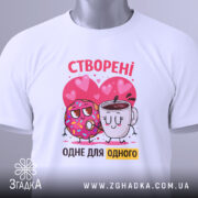 Футболка створені одне для одного. Біла футболка, складена на столі, з принтом, де пончик і чашка з кавою обіймаються на фоні сердечок. Яскраві кольори принта, видно етикетку з логотипом.