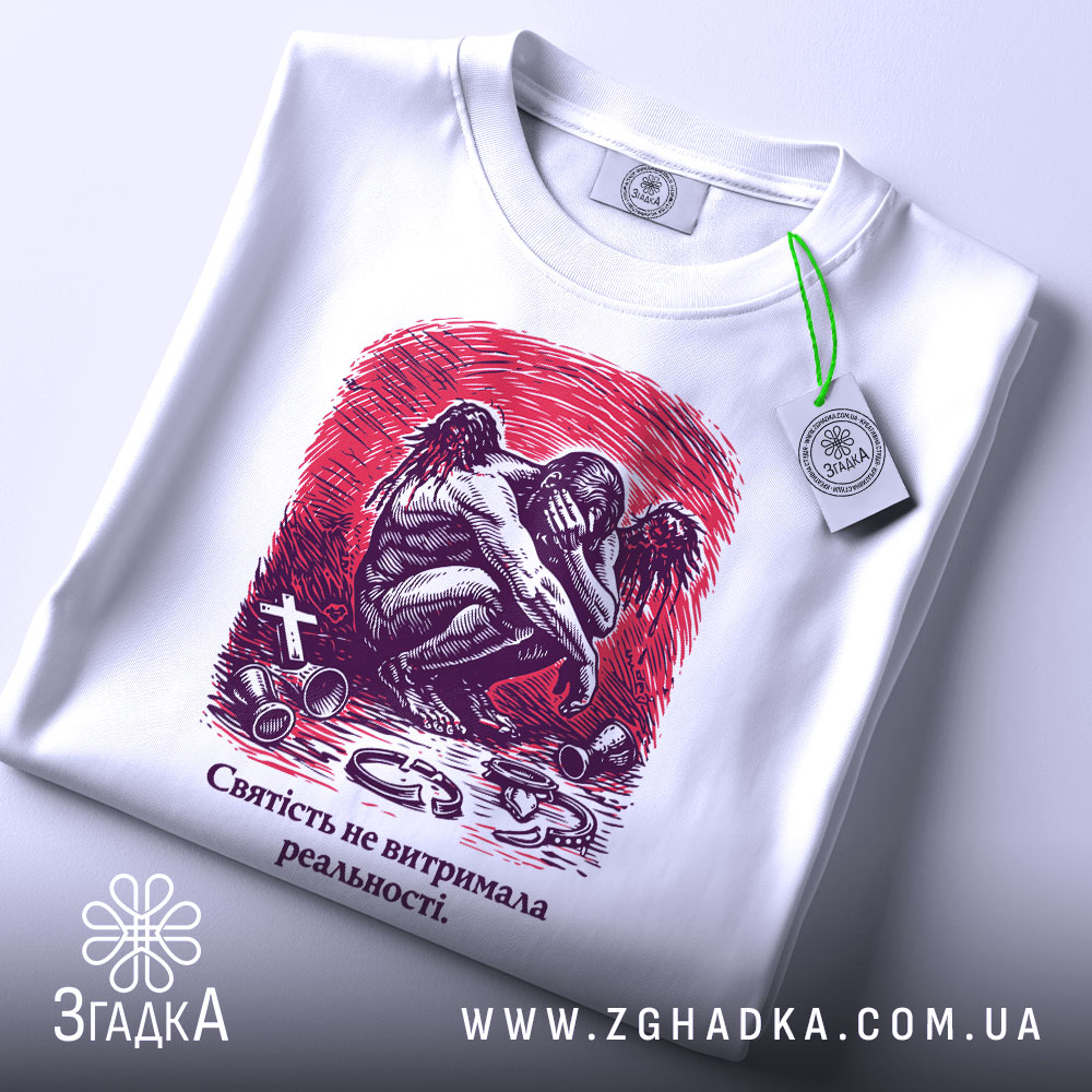 Футболка святість і реальність. Біла футболка з коротким рукавом, зображена з верхнього ракурсу на фоні світлого кольору. На футболці принт з зображенням ангела, що сидить, з червоним фоном. На футболці також видно етикетку з логотипом бренду.