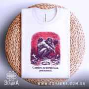 Футболка святість і реальність. Біла футболка, складена на плетеній основі, з нейтральним фоном. Принт з зображенням ангела, що сидить, на червоному фоні, з текстом: 'Святість не витримала реальності.' Видно етикетку з логотипом бренду.