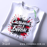 Футболка сильний з Богом, біла, складена на поверхні, фон світло-сірий, принт з червоними та чорними краплями фарби навколо напису 'Сильний, бо з Богом', з етикеткою на зеленому шнурку.