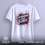 Футболка сильний з Богом, біла, підвішена на вішалці, фон світло-сірий, принт з червоними та чорними краплями фарби навколо напису 'Сильний, бо з Богом'.