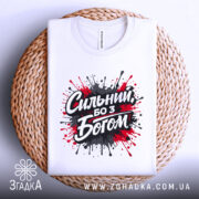 Футболка сильний з Богом, біла, складена на плетеній основі, фон світло-сірий, принт з червоними та чорними краплями фарби навколо напису 'Сильний, бо з Богом'.