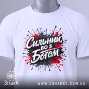 Футболка сильний з Богом, біла, складена на поверхні, фон світло-сірий, принт з червоними та чорними краплями фарби навколо напису 'Сильний, бо з Богом', з етикеткою на зеленому шнурку.