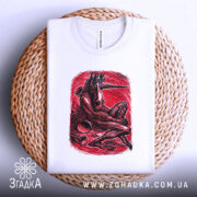 Футболка темний сюрреалізм, біла, складена на плетеній кошику, фон світло-сірий, принт червоного кольору з зображенням істоти з рогами, що сидить, з чіткими деталями принта.